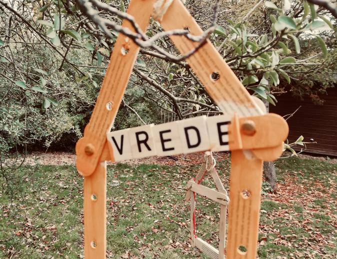 Een ééndimensionaal huisje van houten ijslollystokjes hangt buiten aan een tak. De dwarsbalk onder het dak leest VREDE.