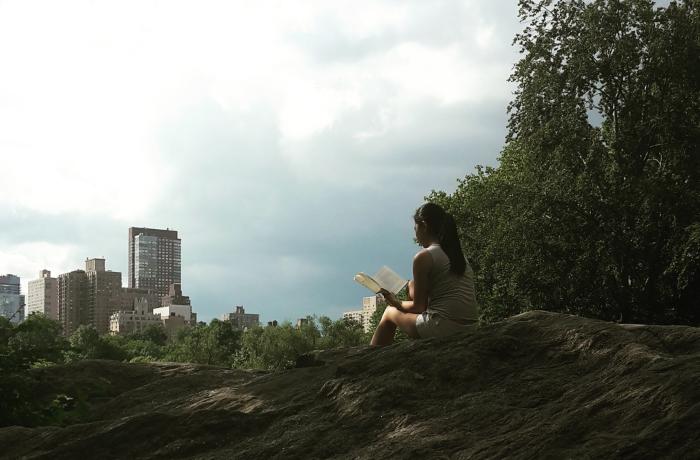 Meisje leest een boek op een rots in het groen van Central Park, New York. Hoge appartementsgebouwen vormen de horizon.